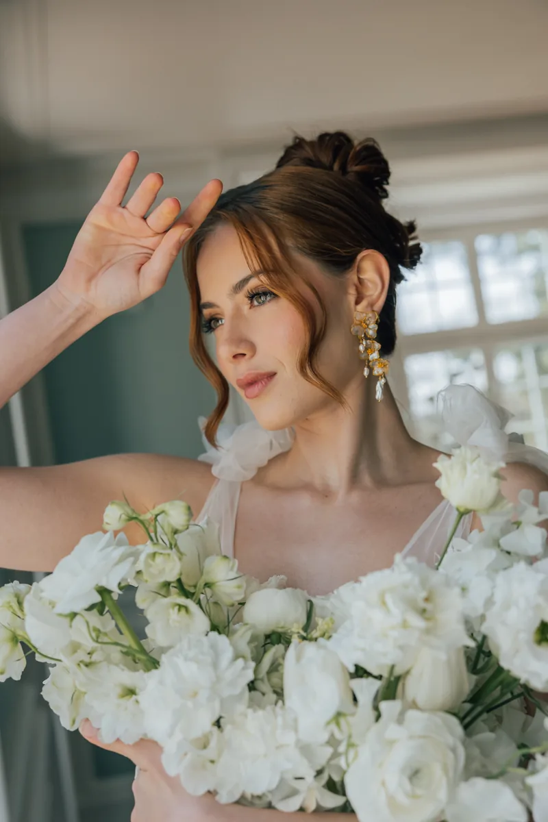 Sposa con chignon, orecchini floreali dorati e un bouquet bianco