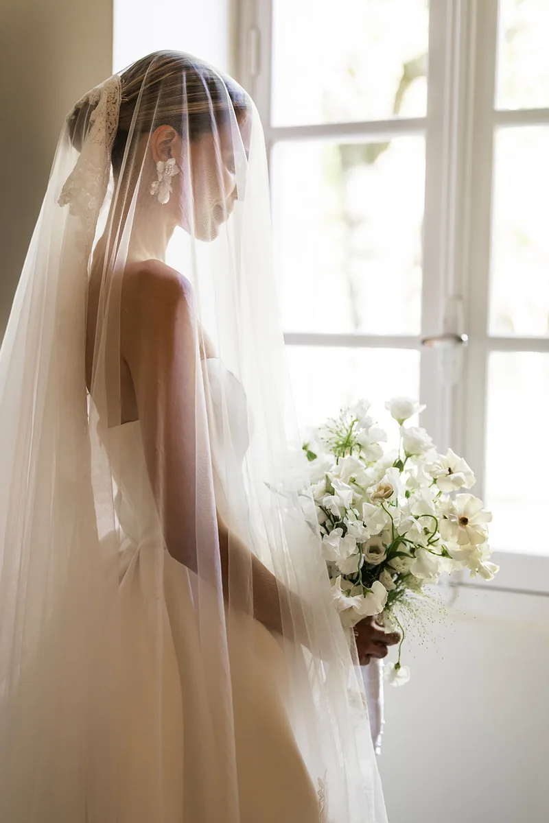 White floral earrings worn under a veil, by a window
